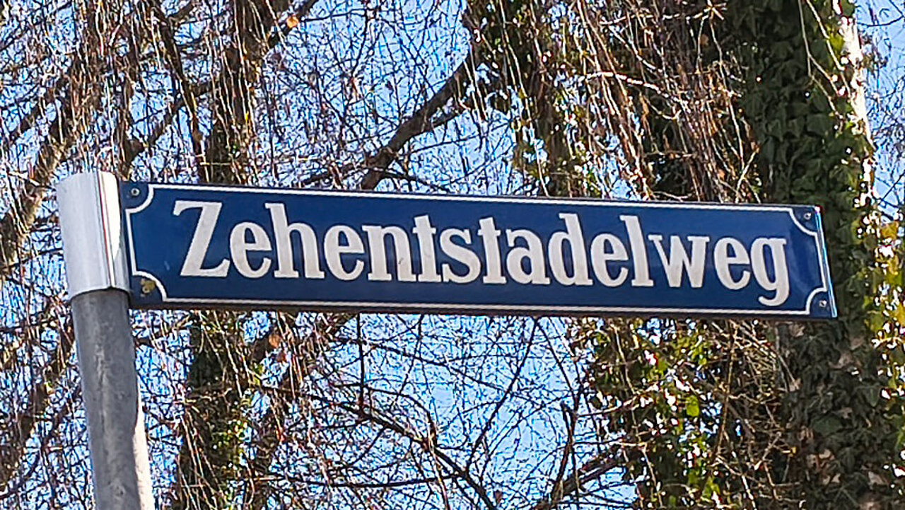 Blaues Straßenschild mit der Aufschrift „Zehentstadelweg“ vor kahlen Bäumen und blauem Himmel.