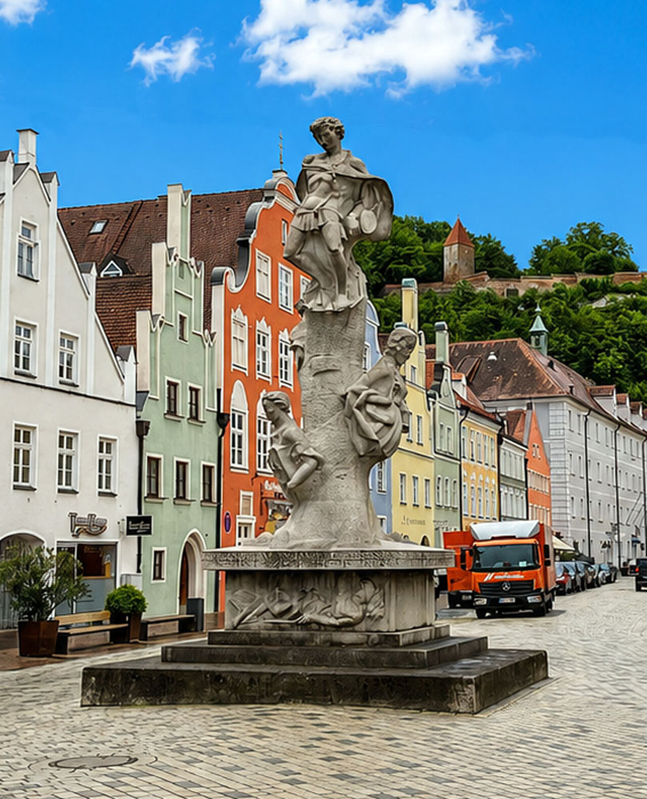 Denkmal auf dem Landshuter Hauptplatz mit historischen Bürgerhäusern und der Burg Trausnitz im Hintergrund.