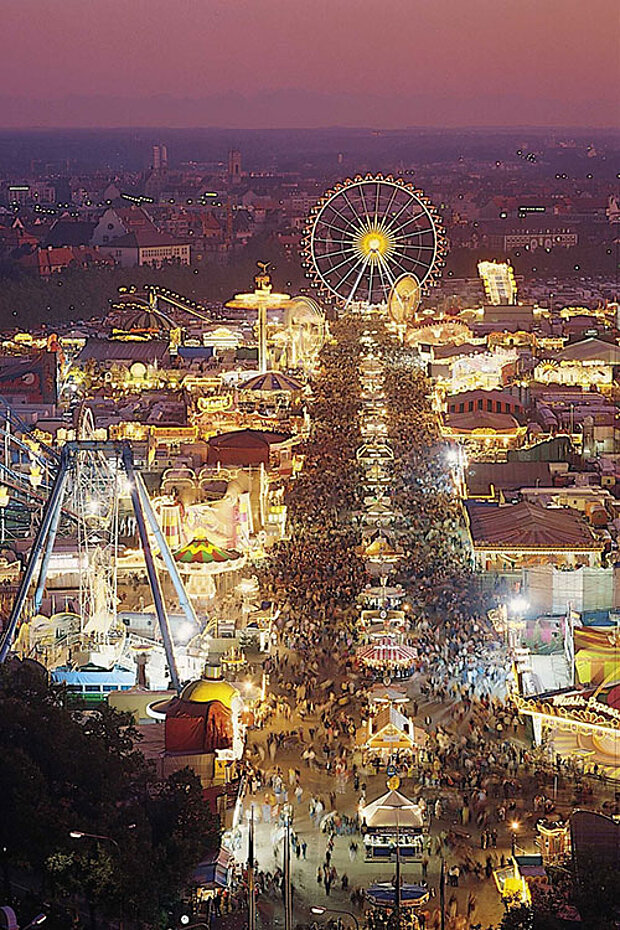 Panorama des Münchner Oktoberfests bei Nacht mit Riesenrad, Fahrgeschäften und vielen Besucherinnen und Besuchern.