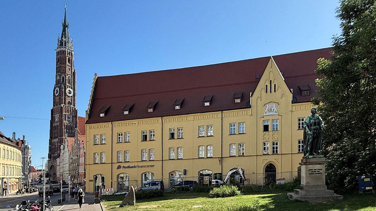 Der Dreifaltigkeitsplatz in Landshut mit historischem Gebäude, Denkmal und dem Turm der Kirche St. Martin im Hintergrund.
