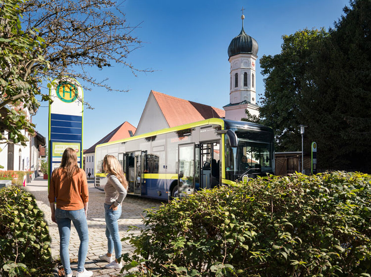 An MVV regional bus at a bus stop in a village. In the background are a church with an onion dome and other buildings; in the foreground is the bus stop.
