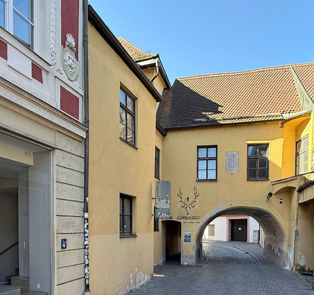 Aufnahme eines historischen Gebäudekomplexes in der Landshuter Altstadt. Ein gewölbter Torbogen führt durch das Gebäude in eine schmale, gepflasterte Gasse. Die Fassaden sind in Gelb- und Cremetönen gehalten, über dem Durchgang befindet sich eine Gedenk- oder Informationstafel.