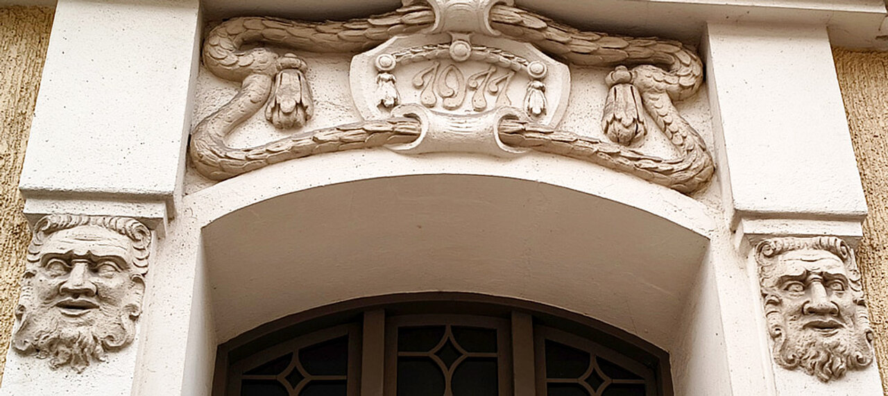 Historischer Hauseingang mit ornamentalem Stuckdekor. Über dem Torbogen befindet sich die Jahreszahl 1911, seitlich sind zwei steinerne Maskenköpfe dargestellt.