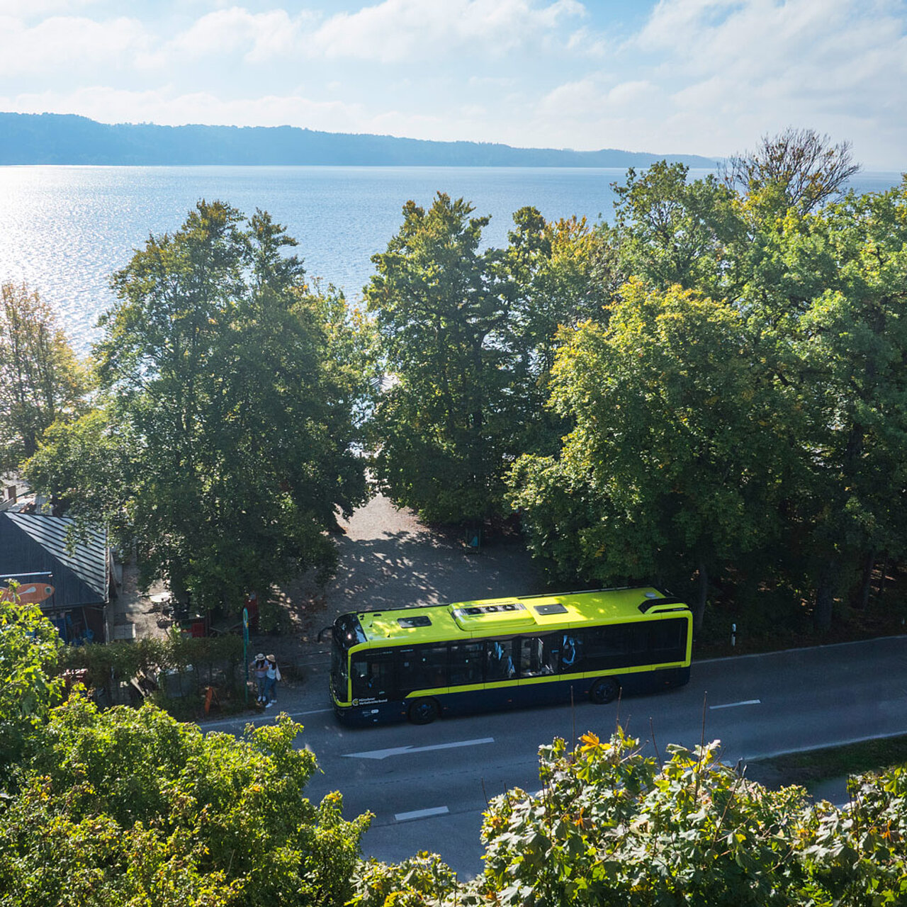 An MVV regional bus on a road running alongside Lake Starnberg. The photograph shows the bus from an elevated vantage point, surrounded by trees, with a view of the lake in the background.