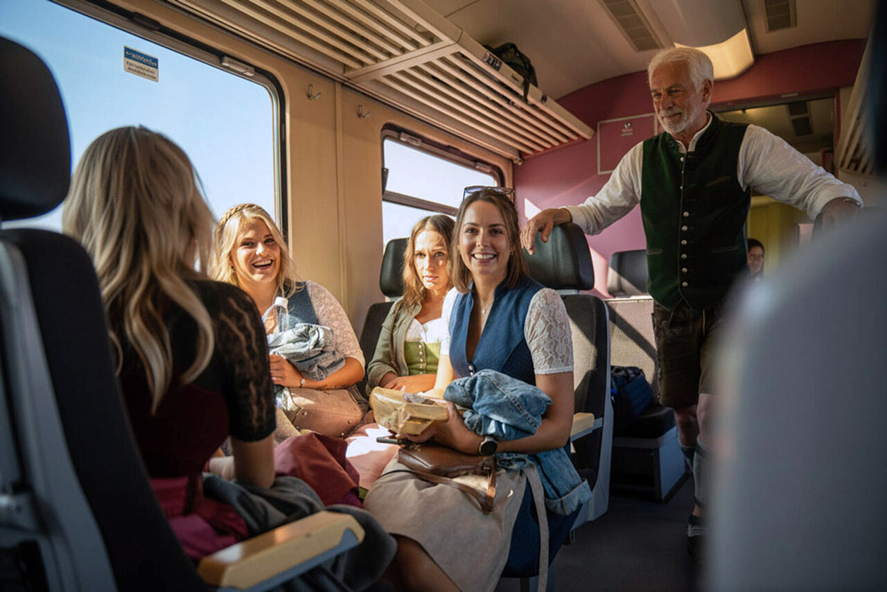 Fahrgäste in traditioneller Festkleidung sitzen in einem Regionalzug und nutzen die Anreise mit öffentlichen Verkehrsmitteln.