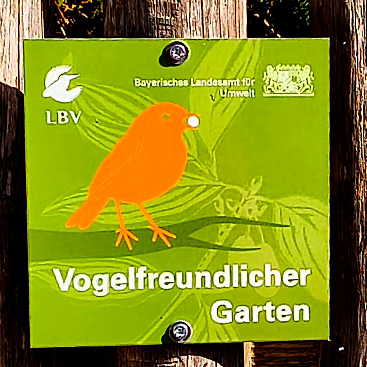 Auszeichnungsschild „Vogelfreundlicher Garten“ an einem Holzzaun. Das grüne Schild zeigt einen stilisierten Vogel und trägt die Logos des Landesbunds für Vogel- und Naturschutz (LBV) sowie des Bayerischen Landesamts für Umwelt.