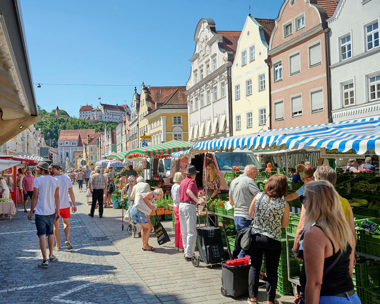 Wochenmarkt in der Landshuter Altstadt mit Marktständen, historischen Bürgerhäusern und der Burg Trausnitz auf dem Hügel über der Stadt.