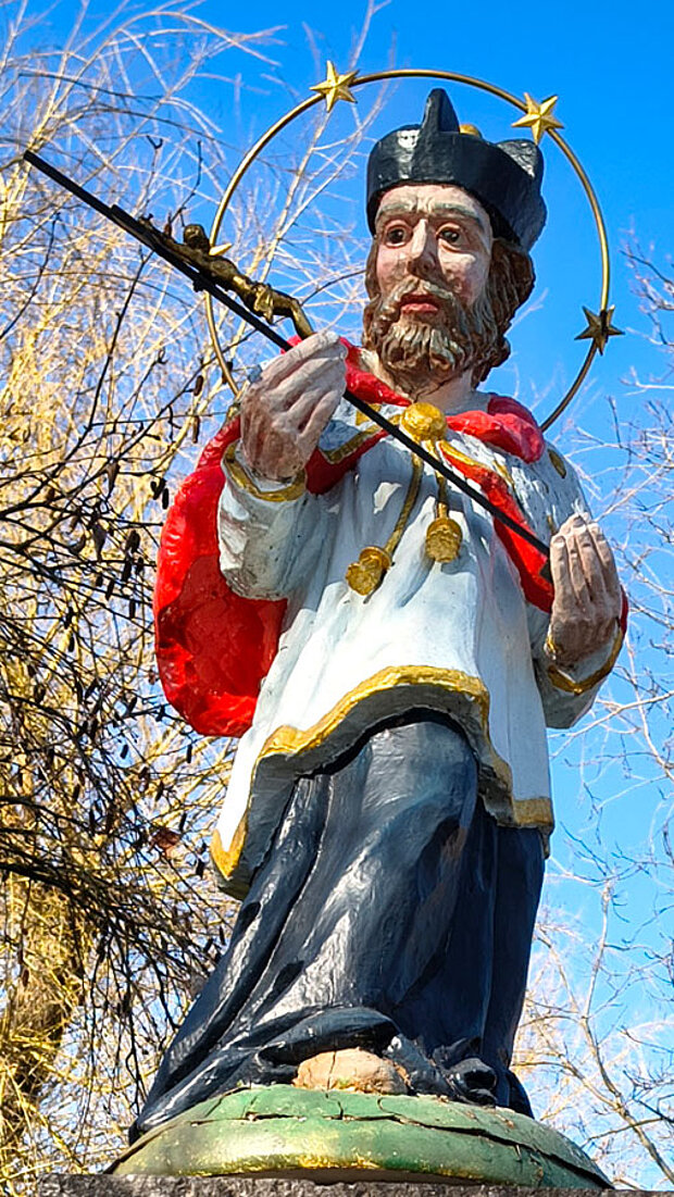 Historische Heiligenfigur im Freien mit rotem Umhang, weißem Gewand und schwarzer Kopfbedeckung. Hinter dem Kopf befindet sich ein goldener Sternenkranz, in den Händen hält die Figur einen langen Stab.