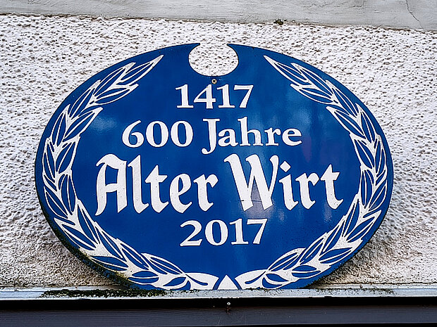 Ovale blaue Jubiläumstafel zur 600-jährigen Geschichte des Alten Wirts. Die Tafel trägt die Aufschrift „1417 – 600 Jahre Alter Wirt – 2017“ und ist von einem stilisierten Lorbeerkranz umrahmt.