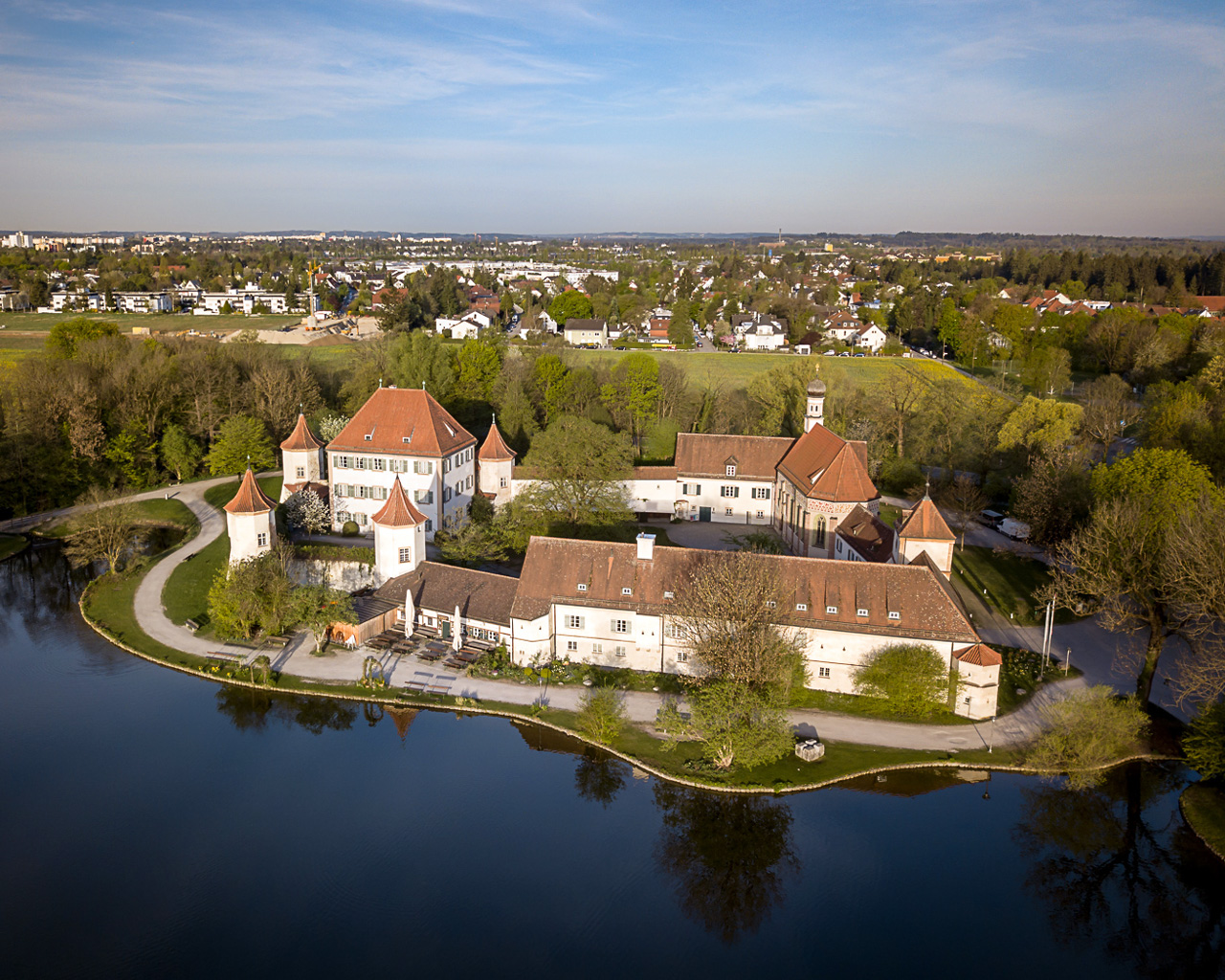Luftaufnahme von Schloss Blutenburg in München-Obermenzing. Die historische Schlossanlage mit mehreren Gebäuden und charakteristischen Türmen liegt auf einer von Wasser umgebenen Insel. Im Hintergrund sind Wohngebiete und Grünflächen zu sehen.